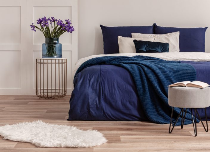 Purple flowers in blue glass vase on stylish bedside table next to king size bed